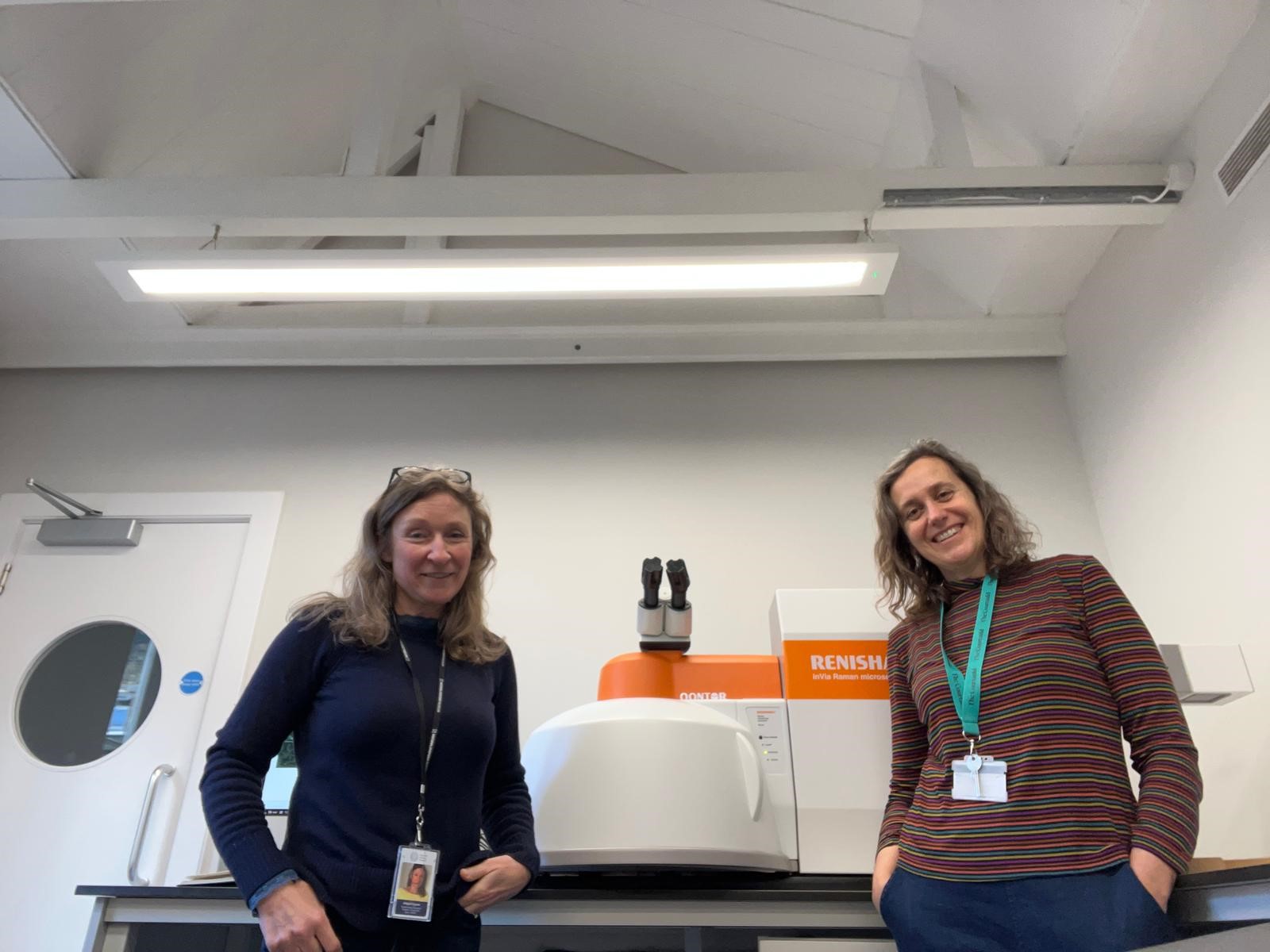 Two smiling women standing side by side a piece of research equipment.